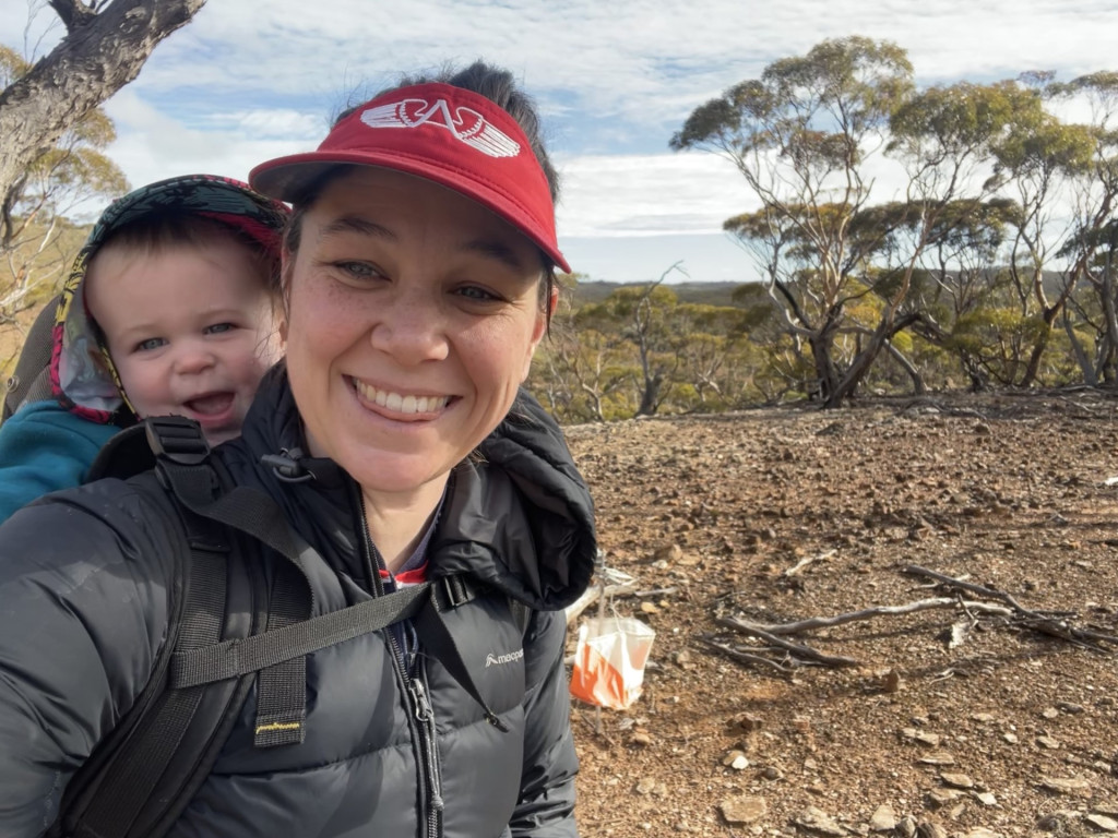 Photo of Bridget with a child on her back in an Australian bush with an orienteering flag